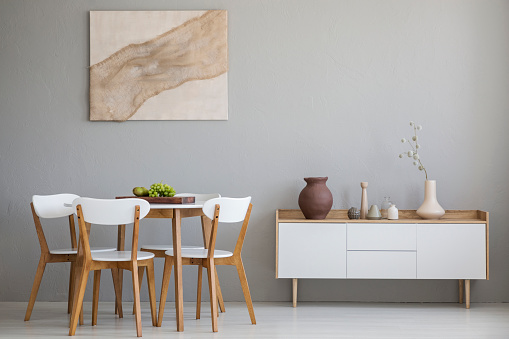Real photo of wooden table and four chairs standing in light grey dining room interior with modern art poster and cupboard with decor