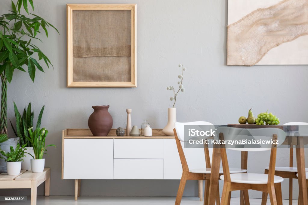 Dining table with fresh fruits and white wooden chairs in real photo of light grey room interior with two posters, green plants and small cupboard with decor Indoors Stock Photo
