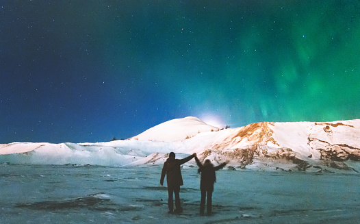 Couple Travelers enjoying Northern lights view above mountains raised hands Travel Lifestyle and relationship man and woman concept vacations into the wild night scene