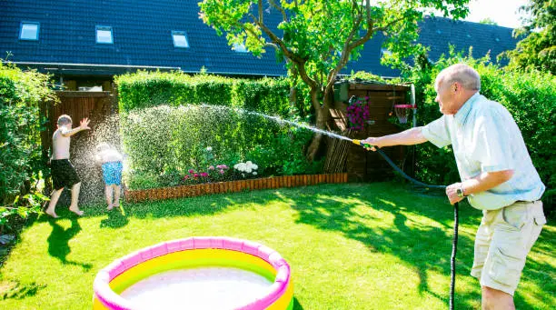 Grandfather uses a garden hose to spray water on his grandkids during a big water fight in a back yard. It is summer and the sun shines.
