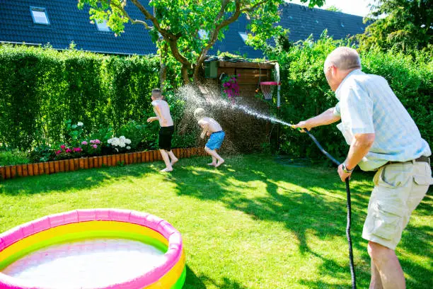 Grandfather uses a garden hose to spray water on his grandkids during a big water fight in a back yard. It is summer and the sun shines.