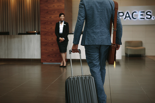 Rear view of businessman arriving the hotel with his luggage with female concierge standing in background for welcoming.