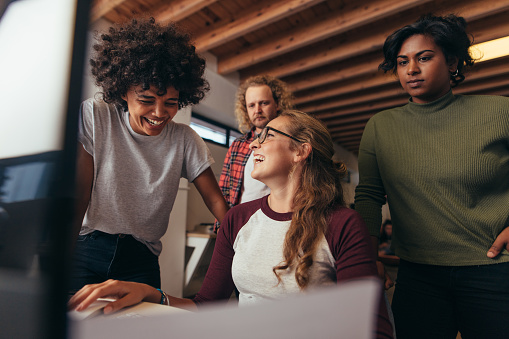 Multi-ethnic startup business team working together in office. Woman sitting at her desk laughing with team standing by. Business team having casual talk while working.