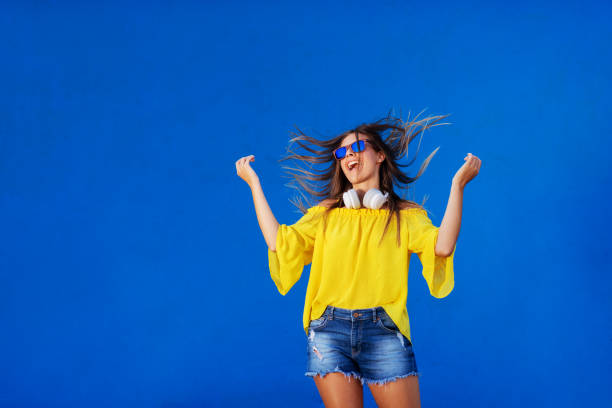 joyeuse fille debout chemise jaune contre le mur bleu avec casque autour du cou. danse et souriant. - female likeness audio photos et images de collection