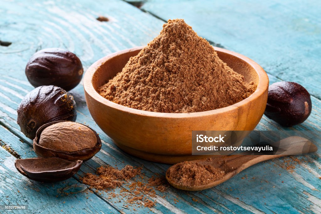 Whole inshell nut, cracked and nutmeg powder in a wooden bowl and spoon on old blue rustic background Whole inshell nut, cracked and nutmeg powder in a wooden bowl and spoon on old blue rustic background close-up. indian spice. Nutmeg Stock Photo