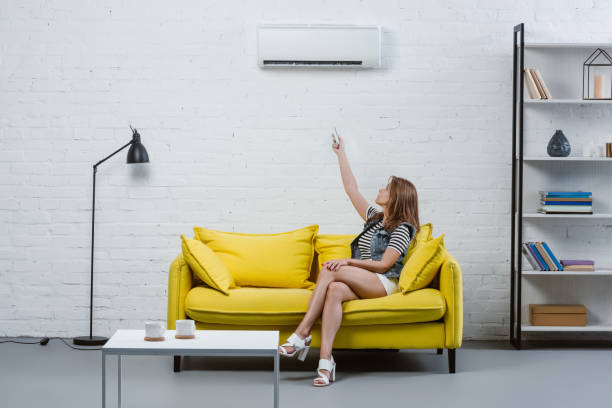 istock attractive young woman sitting on sofa and pointing at air conditioner with remote control 1032763072