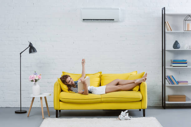 istock relaxed young woman reading book on sofa and pointing at air conditioner with remote control 1032763100