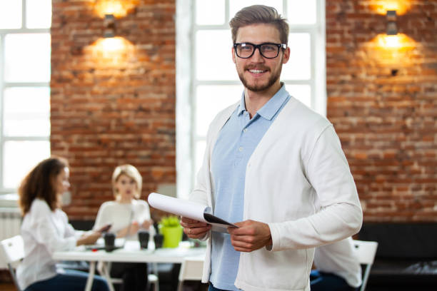 istock Confident team leader. Confident young man keeping arms crossed and looking at camera with smile while his colleagues working in the background. 1032779944