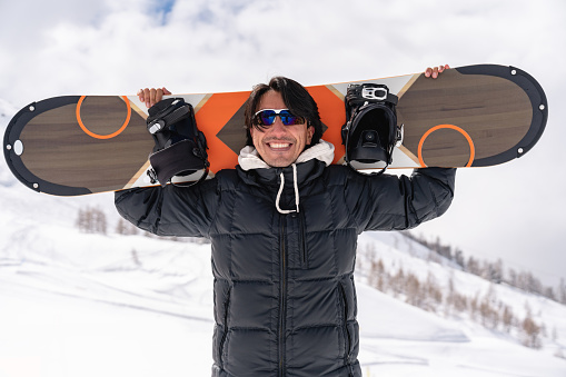 Portrait of a happy snowboarder carrying his board and looking at the camera smiling and wearing goggles - lifestyle concepts