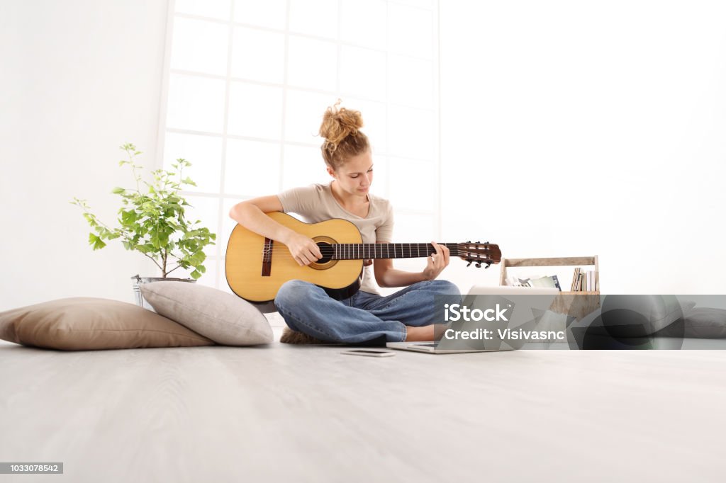 Jouer de la guitare belle jeune femme avec ordinateur, apprendre à jouer avec un cours en ligne, assis sur le sol dans des vêtements décontractés à la maison - Photo de Guitare libre de droits