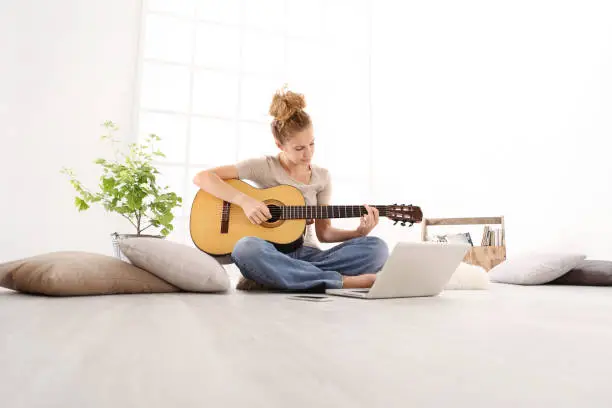 Photo of Beautiful young woman playing guitar with computer, learn to play with an online course, sitting on the floor in casual clothes at home