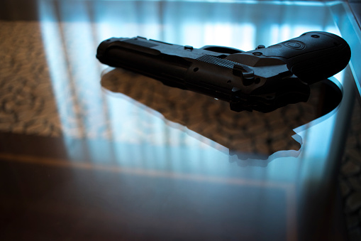 Automatic pistol gun on glass table in bedroom in luxury hotel in silhouette with reflection of window light.