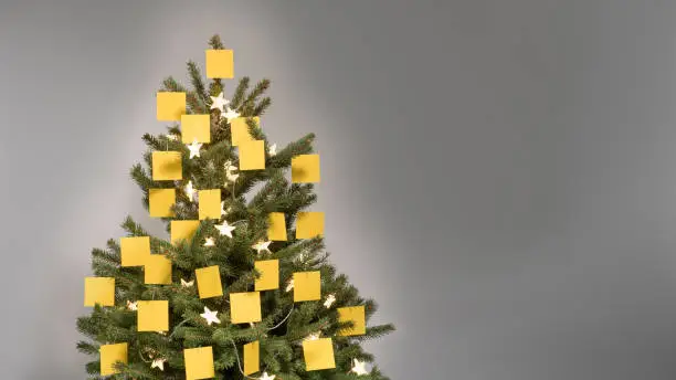 Christmas tree with decorations of 25 blank yellow post-it notes with copy space and fairy lights in front of a light gray background, copy space, nobody