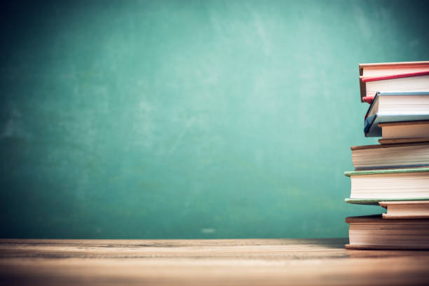 Back to school. Textbooks stacked on wooden school desk in front of green chalkboard. Classroom setting.