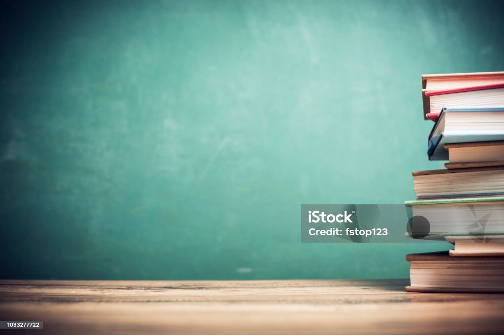 Textbooks on wooden school desk with chalkboard. Back to school.  Textbooks stacked on wooden school desk in front of green chalkboard.  Classroom setting. Education Stock Photo