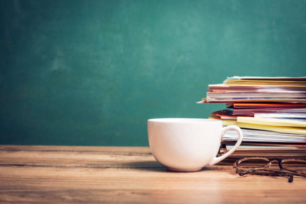 Back to school. Coffee cup on teacher's desk with stack of student papers in front of green chalkboard.  Classroom setting.