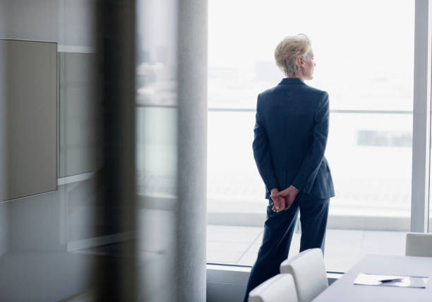 femme d'affaires debout à la fenêtre dans le bureau - regarder par la fenêtre photos et images de collection