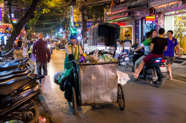 Hanoi, Vietnam, May 11th, 2016. A council rubbish collector pushes her barrow through the streets of the Old Quarter of Hanoi. 8pm with tourists around enjoying the night.