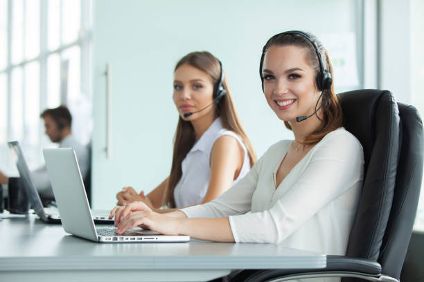 istock Beautiful business people in headsets are using computers and smiling while working in office. Girl is looking at camera. 1033382002