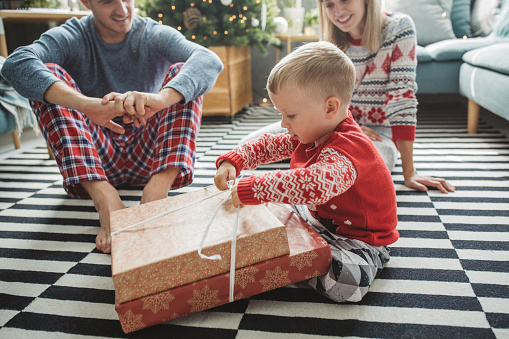 Young parents with baby boy celebrating Christmas at home, boy opening presents . Home is decorated with Christmas ornaments and lights.