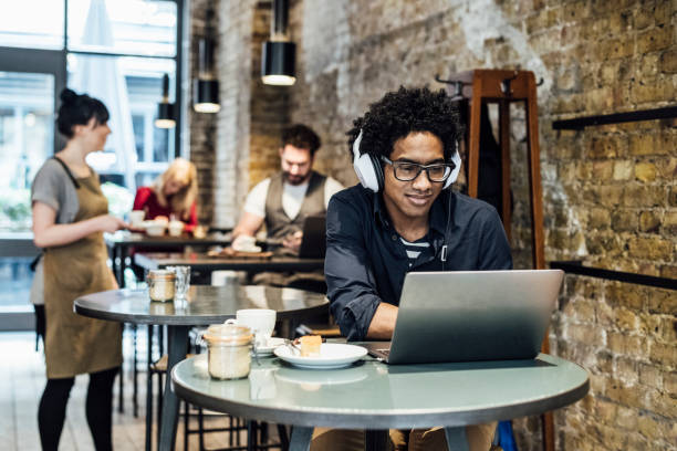 Young man using laptop in cafe and listening to music Male customer sitting at table wearing headphones, waitress serving people in the background coffee shop stock pictures, royalty-free photos & images