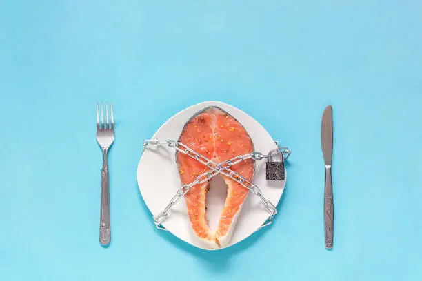 Piece of red fish on plate and chain with closed padlock, fork table-knife on blue background. Concept-prohibition, embargo, danger artificially grown fish or allergy Copy space Flat Lay