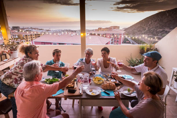 grupo de amigos e pais em casa jantando juntos e tilintar de copos com bebidas. todo mundo sorri e divirta-se a celebrar em amizade. pessoas felizes de caucasianas no terraço em casa com edifícios e vista para o mar durante o pôr do sol - foodie - fotografias e filmes do acervo