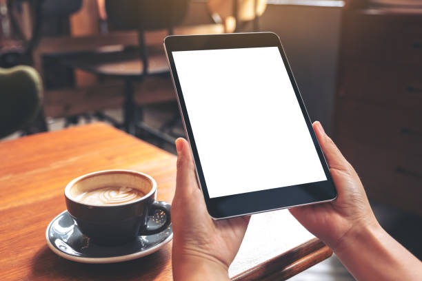 istock Mockup image of hands holding black tablet pc with blank white screen with coffee cup on wooden table in cafe 1033716740