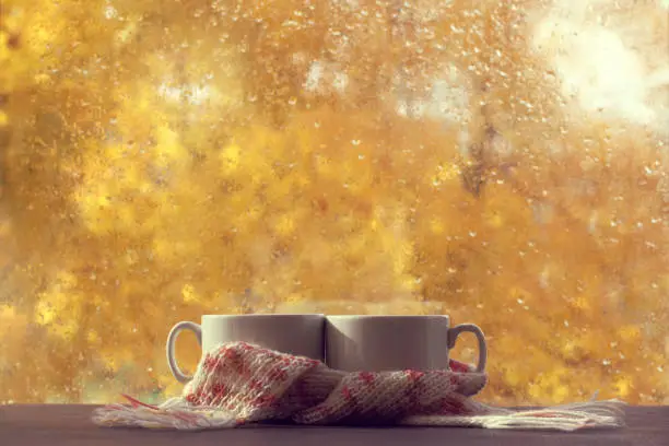 Two white cups in pink scarf on table in front of window with raindrops autumn