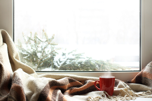 red mug of hot drink on a windowsill with a warm blanket, when behind is window is snow