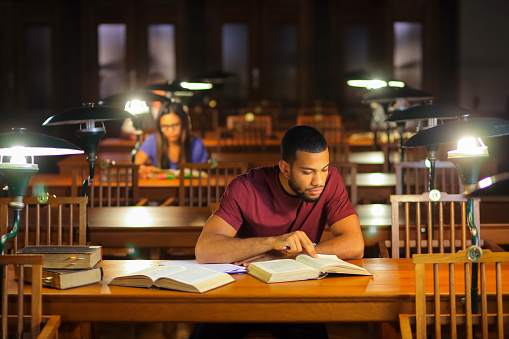 People reading books in the library. Afro-american man and asian woman sitting in the library reading room and studying for exam late at nigh.