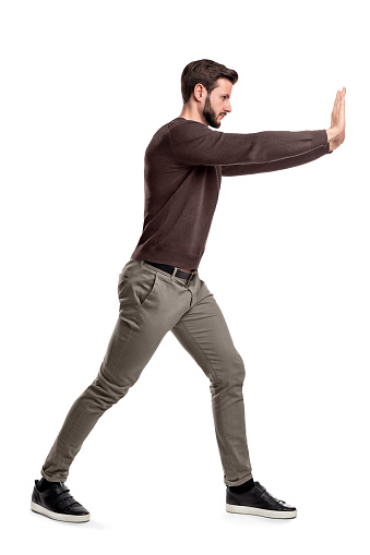 Un hombre con barba en ropa casual trata de empujar un objeto pesado con ambos brazos con una pierna puesta en frente de equilibrio. photo