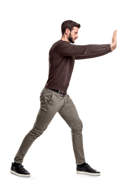 un uomo barbuto in abiti casual cerca di spingere un oggetto pesante con entrambe le braccia con una gamba messa davanti per l'equilibrio. - spingere foto e immagini stock