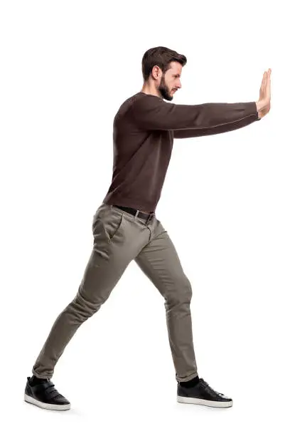 Photo of A bearded man in casual clothes tries to push a heavy object with both arms with one leg put in front for balance.