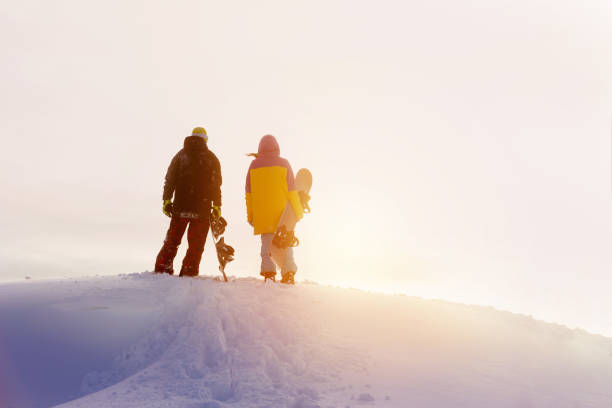 Two snowboarders male and female stands on mountain top and looks at sunset