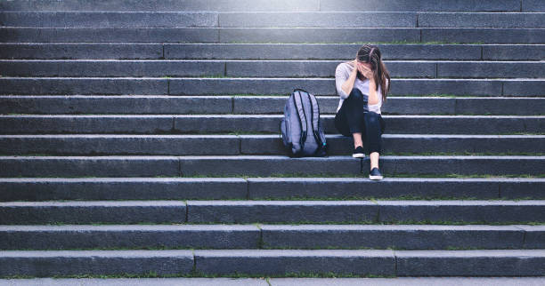 Bullying, discrimination or stress concept. Sad teenager crying in school yard. Upset young female student having anxiety. Upset victim of abuse or harassment sitting on stairs outdoors. Bullying, discrimination or stress concept. Sad teenager crying in school yard. Upset young female student having anxiety. Upset victim of abuse or harassment sitting on stairs outdoors with backbag. teasing photos stock pictures, royalty-free photos & images