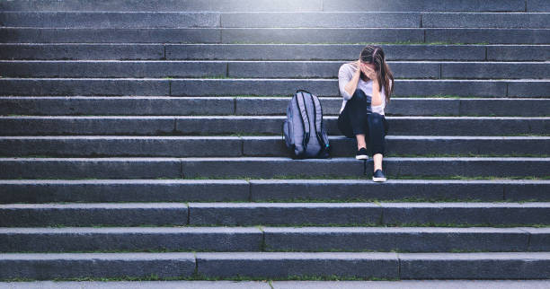 zastraszanie, dyskryminacja lub stres. smutny nastolatek płacze na szkolnym podwórku. zdenerwowany młoda studentka o lęku. zdenerwowany ofiara nadużyć lub molestowania siedzi na schodach na zewnątrz. - exclude zdjęcia i obrazy z banku zdjęć