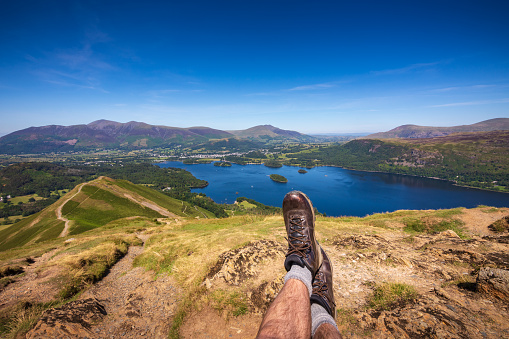 A lone male hiker is sitting down and resting his feet while he enjoys the spectacular views from the Cat Bells fell. Down below is the beautiful tranquil scene of Lake Derwentwater and the town of Keswick in the English Lake District. His hiking boots are well worn and broken in.