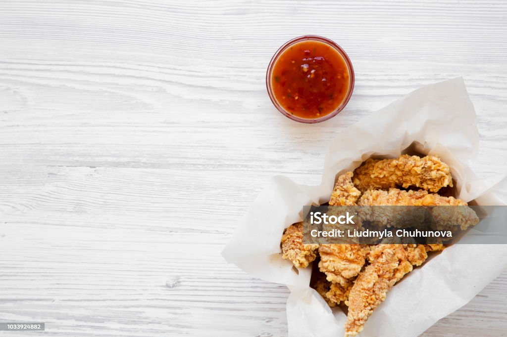 Strisce di pollo in scatola di carta con salsa al peperoncino dolce su un tavolo di legno bianco, vista dall'alto. Lay piatto, vista dall'alto, sopra la testa. Copiare lo spazio. - Foto stock royalty-free di Carne di pollo