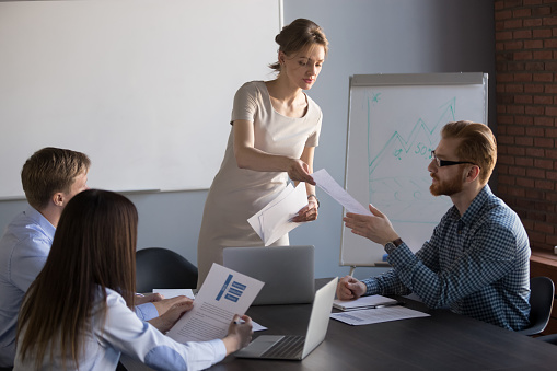 Millennial businesswoman give handout materials to work team members during flipchart presentation, female speaker or coach share documents to workers, presenter hand papers to consider or analyze