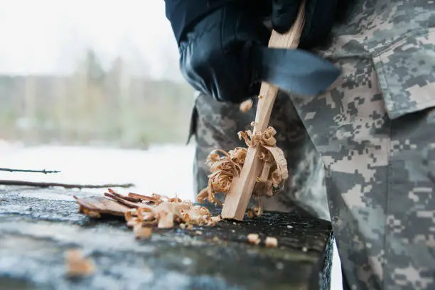Man prepares wood-wool with hunting knife for starting fire. Wood shaving. Survival concept