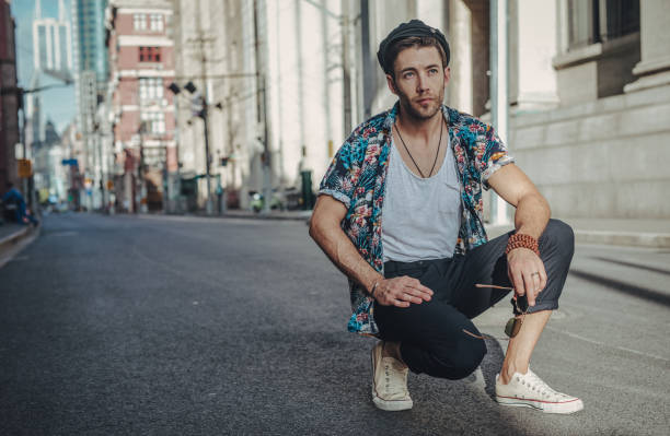 Young hipster man in black fashionable clothes in trendy sunglasses in stylish white sneakers is sitting on the asphalt in the city. Handsome guy resting on a summer sunny day outdoors. Street style.
