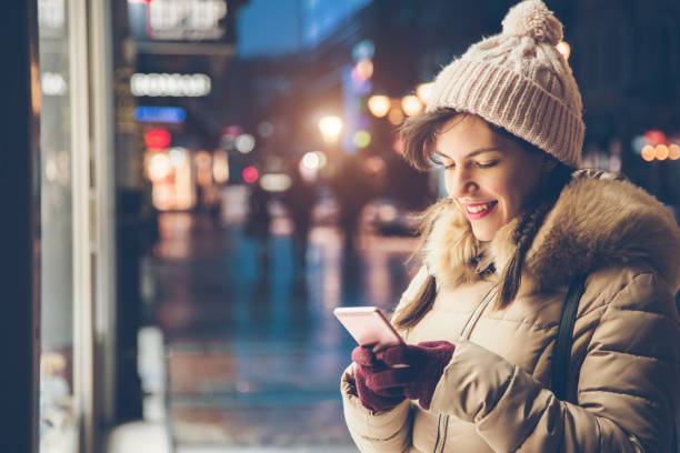 Woman using phone in the winter Young brunette woman typing on her phone in the street during winter window shopping at night stock pictures, royalty-free photos & images