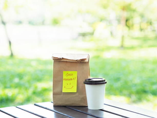 Take away breakfast in the park. Cup of hot coffee and brown paper bag with note Good Morning on wooden table.