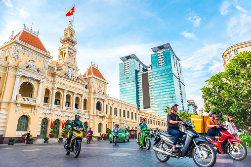 Ho Chi Minh City, Vietnam - April 30, 2018: Saigon City Hall, Vincom Center towers, colorful street traffic (people driving motorbikes) & tropical plants against the blue sky.