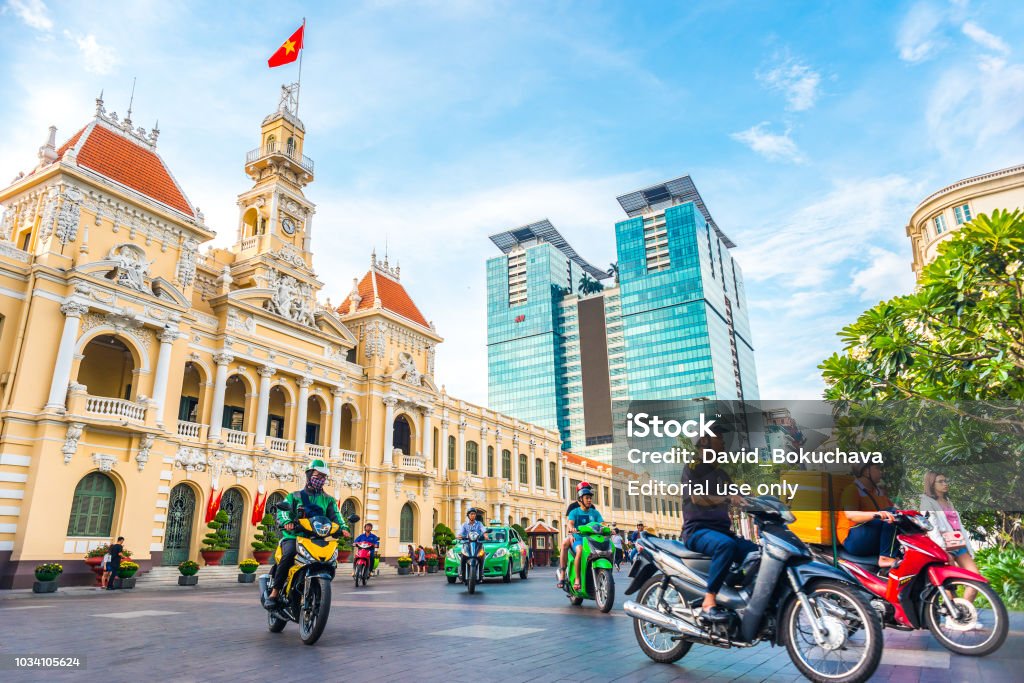 Ho Chi Minh City Ho Chi Minh City, Vietnam - April 30, 2018: Saigon City Hall, Vincom Center towers, colorful street traffic (people driving motorbikes) & tropical plants against the blue sky. Ho Chi Minh City Stock Photo