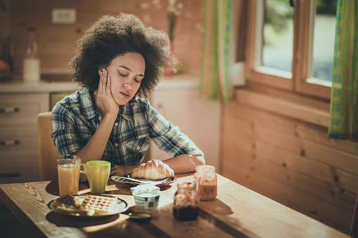 African American woman sitting at dining table and feeling tired to eat her breakfast.