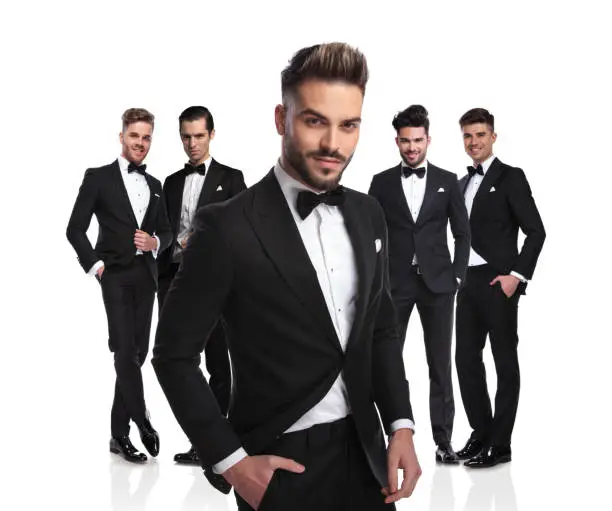 handsome leader of young men in black tuxedoes standing on white background, looking relaxed