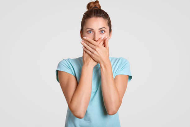 istock Keep silence, please. Frightened woman covers mouth with both palms, looks with scared expression directly at camera, wears casual t shirt, keeps information in secret. Speak no evil concept. 1034211934