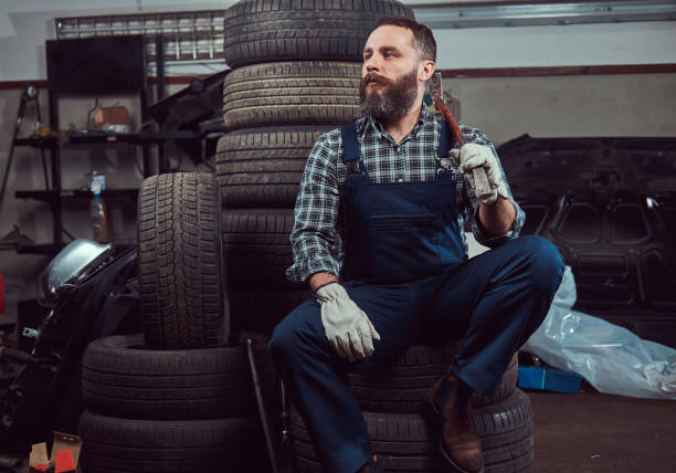 ein bärtiger mechaniker in einer uniform gekleidet sitzt auf alten autoreifen in der garage. - tire auto repair shop part of vehicle stack stock-fotos und bilder